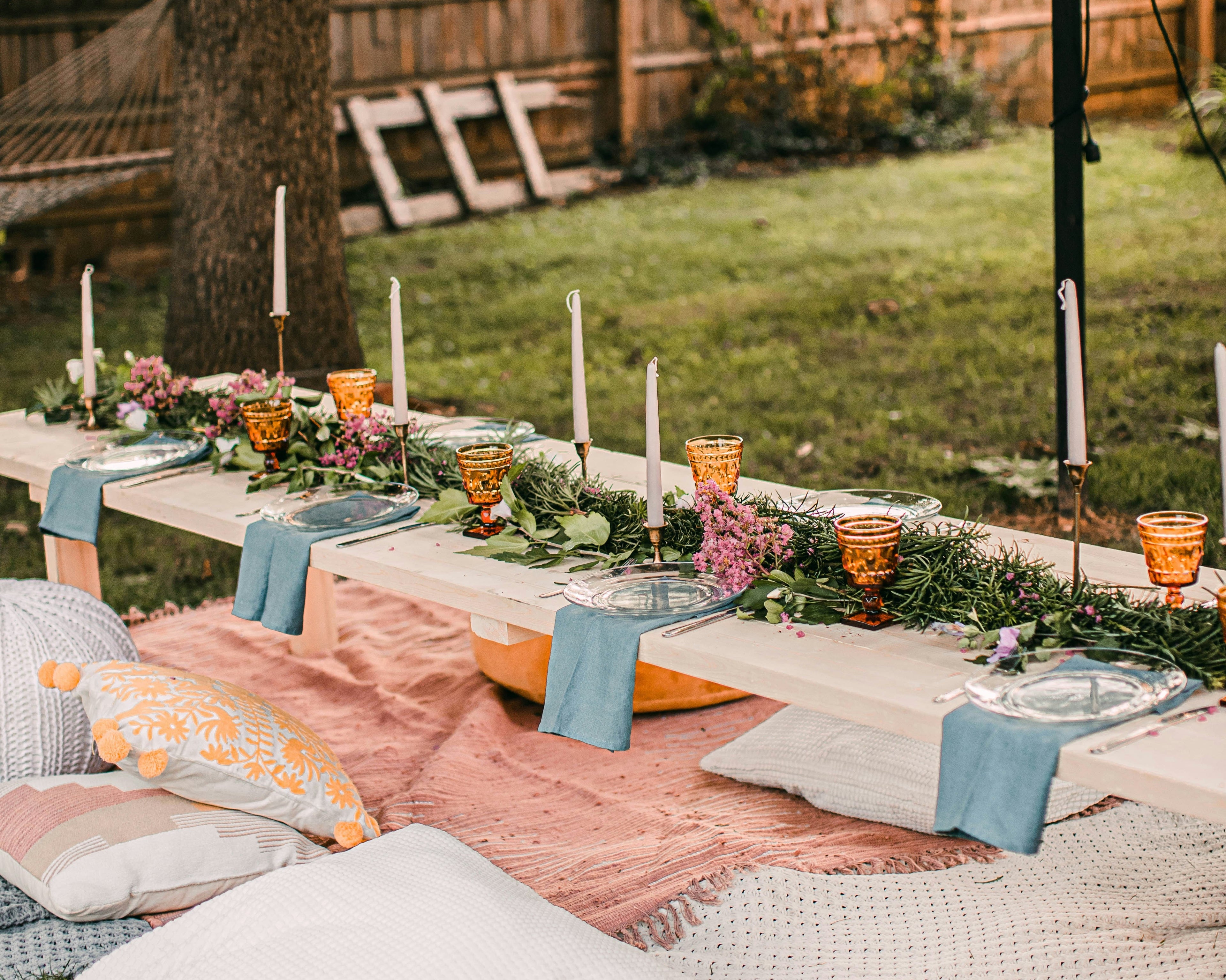 Outdoor setting with a decorated table, candles, and pillows on a grassy area.
